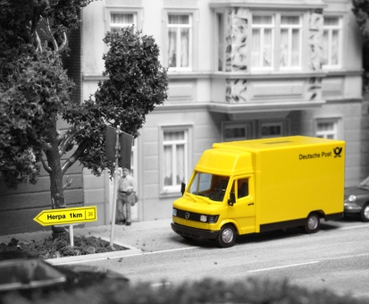 Herpa Available Again, image of a diorama in black and white with yellow accents: Yellow German postal delivery van with yellow sign pointing towards Herpa in a miniature cityscape.